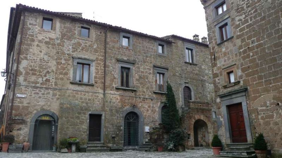 Piazza lastricata in pietra circondata da edifici storici in muratura di pietra, con finestre rettangolari e portali ad arco. Al centro si vedono una piccola scalinata, un arco passante e vasi con piante vicino agli ingressi.