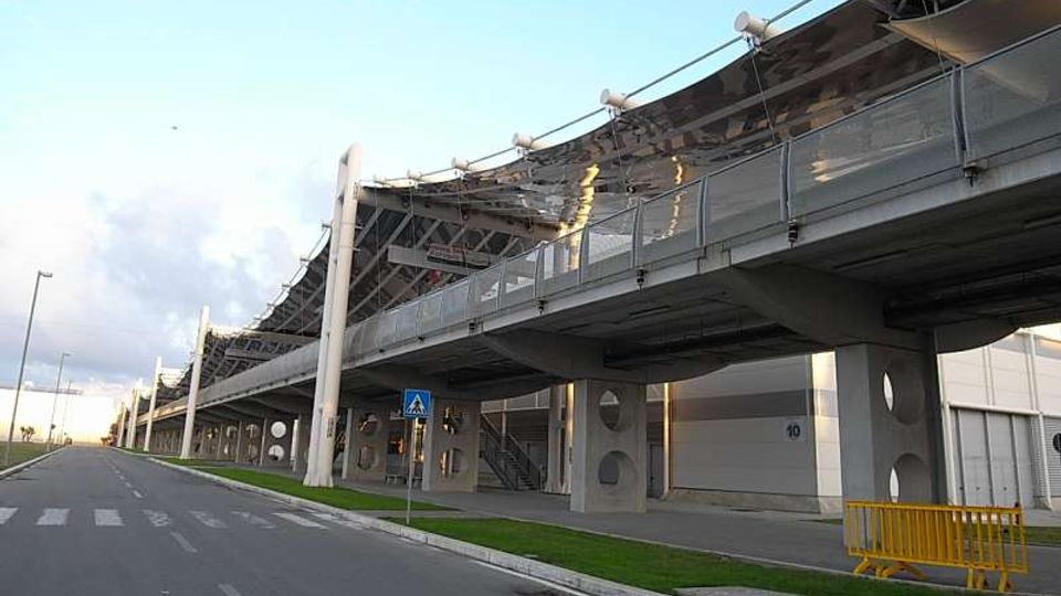 Vista esterna di una grande struttura moderna sopraelevata con copertura curva e passerella con parapetti metallici, sostenuta da pilastri in cemento con aperture circolari. In primo piano si vede una strada asfaltata con marciapiede e striscia di prato, accanto a un edificio adiacente con ingresso numerato.