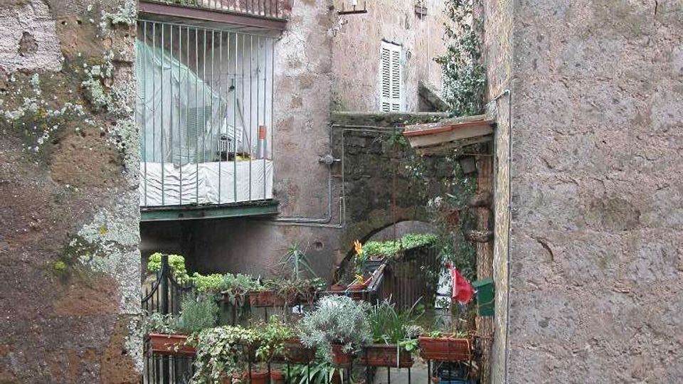 Cortile interno tra edifici in pietra e intonaco con balconi e finestre, visto attraverso un passaggio stretto. In primo piano una ringhiera con numerosi vasi di piante e, sotto, una breve scalinata che scende verso un’area inferiore.