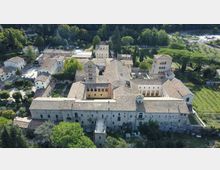 Veduta aerea di un grande complesso monastico in pietra con tetti in coppi, disposto attorno a cortili interni e con una torre campanaria. L’edificio è circondato da alberi e aree verdi, con campi coltivati e parcheggi nelle vicinanze.