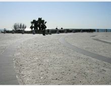 Ampia piazza panoramica pavimentata in pietra chiara con percorsi curvilinei in lastre scure, affacciata sul mare tramite una ringhiera. Sullo sfondo si vedono una palma, alberi spogli e alcune panchine lungo il belvedere.