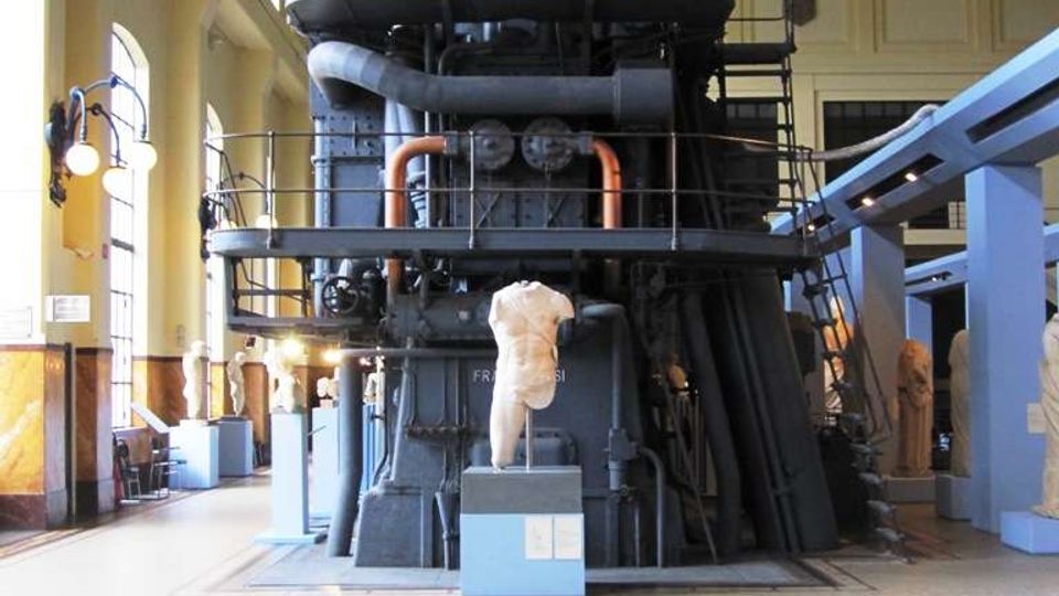 Interno di un museo tecnico in una grande sala industriale con alte finestre ad arco e soffitto a capriate. Al centro si trova un enorme macchinario metallico scuro con tubazioni e passerelle, circondato da pannelli espositivi e basamenti.