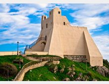 Fortezza in muratura chiara su un promontorio erboso e roccioso, con pareti inclinate, piccole finestre e una torre superiore. Una scalinata con ringhiere e lampioni conduce all’ingresso, sotto un cielo azzurro con nuvole.