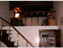 Interno di una casa con soppalco in legno e parapetto con colonnine, raggiungibile tramite una scala in legno con corrimano. In basso si vede una porta aperta su una libreria a muro, con pareti chiare e soffitto con travi scure.