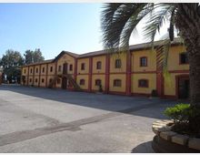 Ampio cortile asfaltato davanti a un lungo edificio a due piani in stile industriale, con facciata gialla e cornici rosse, finestre ad arco e scale esterne. In primo piano si vede una palma e ai lati altre aree verdi con alberi.