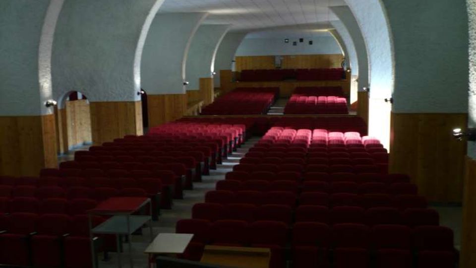 Interno di una sala auditorium con soffitto a volta e grandi archi laterali, pareti chiare e rivestimento in legno lungo i lati. File di poltroncine rosse disposte in platea con corridoio centrale e palco rialzato sul fondo.