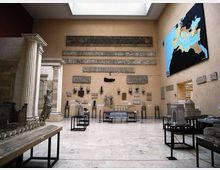 Sala museale con pareti beige e lucernario, con iscrizioni e frammenti lapidei antichi esposti a parete e su pedane. Ai lati si vedono modelli architettonici e reperti in vetrina, e sulla parete destra un grande pannello con una mappa.