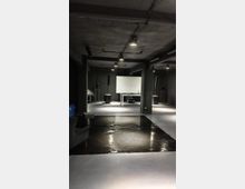Interno di uno spazio industriale scuro con soffitto in cemento a vista e lampade sospese, pareti nere e pavimento lucido. Al centro ci sono un grande schermo bianco su un tavolo e diverse casse acustiche su supporti distribuite nella sala.