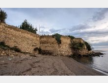 Antiche mura in pietra lungo una spiaggia sabbiosa e ghiaiosa, con vegetazione in cima e tratti erosi vicino alla riva. Il mare calmo si estende sulla destra sotto un cielo nuvoloso.