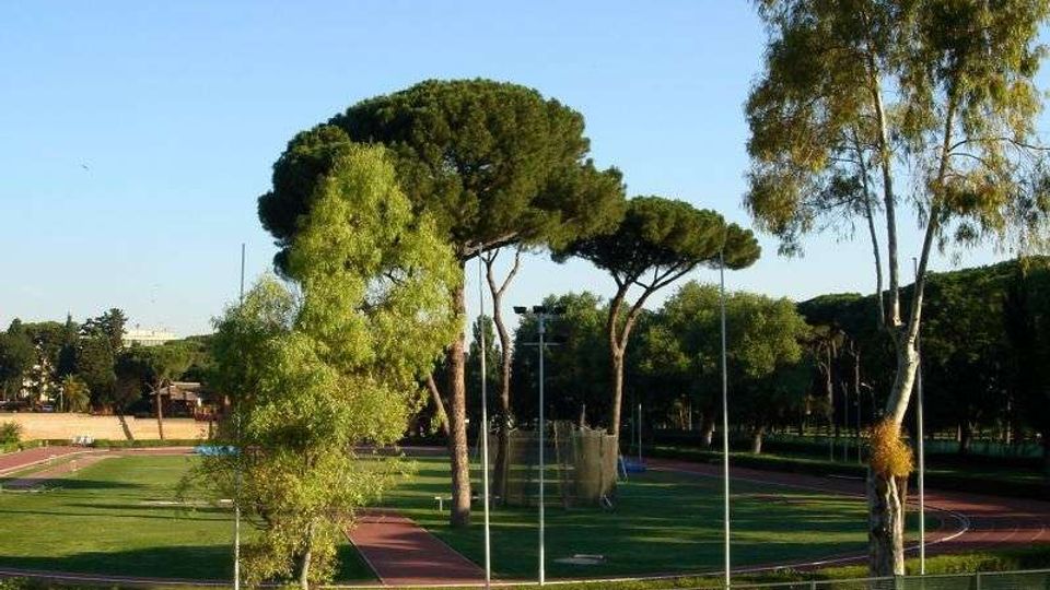 Campo sportivo all’aperto con pista di atletica rossa che circonda un prato, circondato da alte reti e pali per l’illuminazione. Sullo sfondo si vedono alberi ad alto fusto e una linea di vegetazione sotto un cielo sereno.