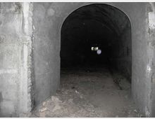 Ingresso ad arco di un tunnel sotterraneo in cemento, con pareti grezze e pavimento coperto di detriti. Il corridoio è buio e si intravede una piccola apertura luminosa in fondo.