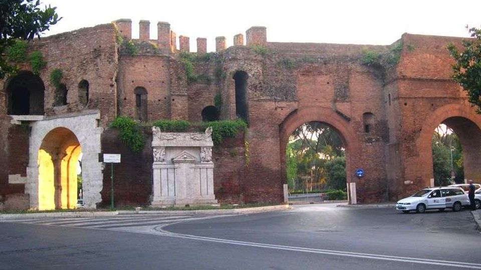Mura antiche in mattoni con merlature e grandi arcate, con edicole in pietra e aperture che conducono a un’area verde retrostante. In primo piano si vede una strada urbana con curva, segnaletica e alcune auto parcheggiate vicino all’arco.