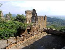 Ruderi di un edificio in pietra su un crinale, con muri parzialmente crollati e vegetazione che cresce intorno. In primo piano c’è una terrazza pavimentata con parapetto in legno e muretto, affacciata su colline boscose.