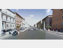 Ampio viale cittadino con carreggiata a più corsie e traffico, fiancheggiato da edifici storici in muratura di diversi piani. Marciapiedi larghi con file di auto in sosta, palme in grandi fioriere e cielo sereno sullo sfondo.