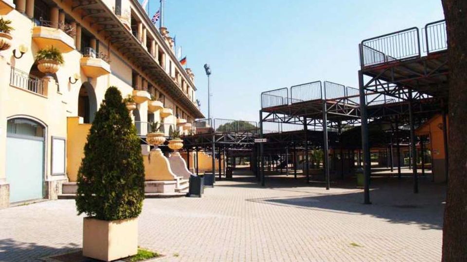 Piazzale pavimentato all’aperto accanto a un edificio a più piani con balconi e arcate, con grandi fioriere e una scalinata d’ingresso. Sul lato destro si vedono strutture metalliche sopraelevate con ringhiere, simili a tribune o passerelle, sotto un cielo sereno.