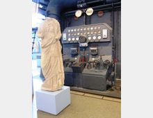 Interno industriale con grande pannello di controllo in metallo, pieno di manometri, leve e volantini, collegato a tubazioni scure. In primo piano, una statua classica senza testa su piedistallo bianco, in uno spazio tipo museo o impianto storico.