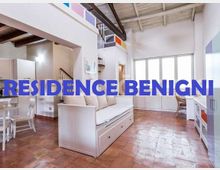 Soggiorno luminoso in open space con pavimento in cotto e soffitto alto con travi a vista, arredato con divano bianco e zona pranzo con tavolo e sedie. Sul fondo ampie porte-finestre vetrate e mobili bianchi lungo la parete.