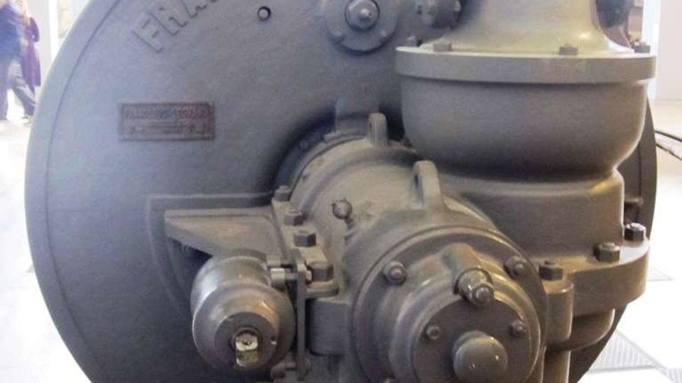 Macchinario industriale d’epoca in metallo grigio, con grande volano circolare e componenti meccaniche a vista, esposto in un ambiente interno tipo museo o spazio espositivo illuminato. Sul volano è visibile la scritta “FRANCO TOSI”.