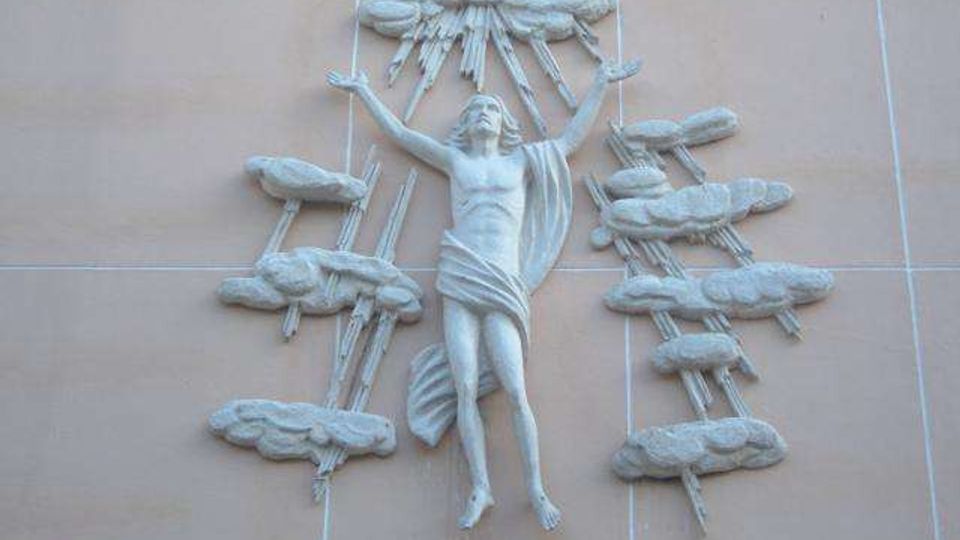 Bassorilievo in pietra su una parete esterna con grandi riquadri, raffigurante una figura centrale con le braccia alzate circondata da nuvole e raggi stilizzati. La scultura è applicata direttamente alla facciata e risalta per il colore chiaro sullo sfondo uniforme.