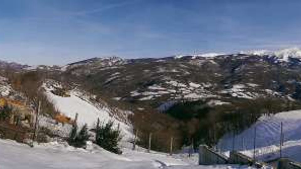 Panorama montano innevato con vallate e crinali sullo sfondo, sotto un cielo limpido. In primo piano si vedono case e recinzioni su un pendio, con neve sui tetti e piccoli alberi sparsi.