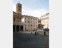 Piazza lastricata in pietra con edifici storici dai toni chiari e una torre campanaria con grande orologio sulla facciata. Al centro si vede una fontana monumentale rialzata con vasca e sculture, circondata da gradini e paletti.