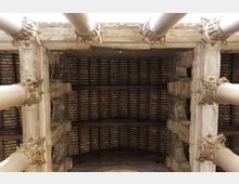 Veduta dal basso del soffitto a cassettoni in legno di un edificio storico, incorniciato da grandi colonne in pietra con capitelli decorati. La struttura mostra travi trasversali e dettagli architettonici in muratura lungo il perimetro.