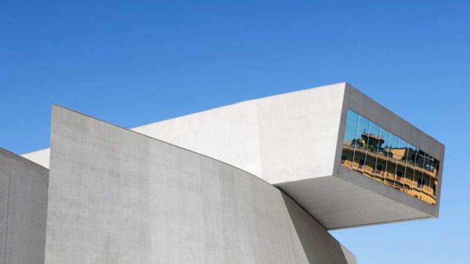 Edificio moderno in cemento con forme geometriche inclinate e un grande volume a sbalzo. All’estremità dello sbalzo c’è una vetrata riflettente, con cielo azzurro sullo sfondo.