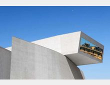 Edificio moderno in cemento con forme geometriche inclinate e un grande volume a sbalzo. All’estremità dello sbalzo c’è una vetrata riflettente, con cielo azzurro sullo sfondo.