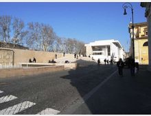 Piazza urbana con ampia scalinata in pietra e piccola fontana a raso sul lato sinistro, affacciata su un edificio moderno bianco con grandi vetrate. Sullo sfondo ci sono alberi spogli e lampioni lungo il marciapiede accanto a edifici storici.