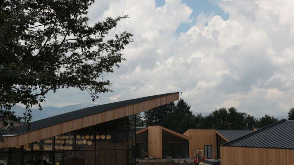 Complesso di edifici moderni in legno con ampie vetrate e tetti inclinati, disposto in più volumi affiancati. In primo piano si vede un’area esterna con sedie e una zona di cantiere, con alberi ai lati e cielo nuvoloso sullo sfondo.