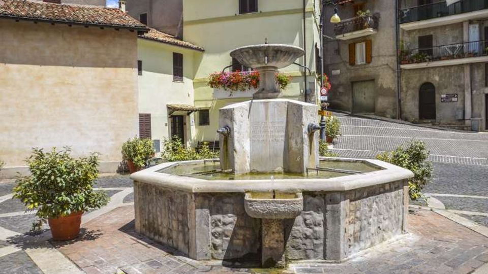 Fontana in pietra al centro di una piccola piazza lastricata, con vasca poligonale e colonna centrale con vaschetta superiore e beccucci da cui scorre l’acqua. Intorno si vedono edifici residenziali a più piani con persiane e balconi, alcuni vasi di piante e una strada in lieve salita sullo sfondo.