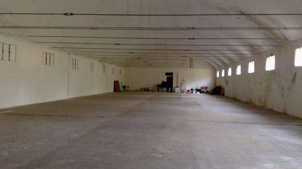 Ampio capannone o magazzino vuoto con pavimento in cemento e soffitto alto attraversato da lunghe travi e cavi. Pareti bianche con una fila di piccole finestre sul lato destro e alcuni materiali accatastati in fondo vicino a una porta.