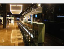 Interno di un moderno bar in stile minimalista, con lungo bancone vetrato e fila di sgabelli in metallo su pavimento lucido. Soffitto con grandi pannelli luminosi e luci a sospensione, pareti scure e area di servizio sullo sfondo.