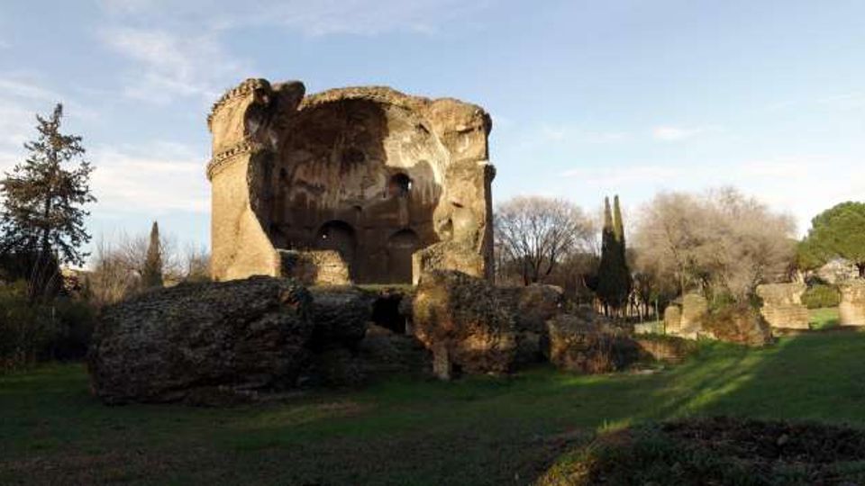 Rovine di una grande struttura circolare in pietra, con ampie parti crollate, situata in un’area erbosa. Intorno si vedono alberi e resti di murature più basse, sotto un cielo sereno.