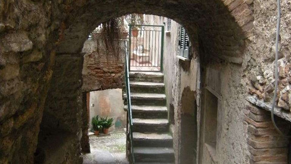 Vicolo stretto in pietra con pavimentazione in ciottoli, attraversato da un arco in mattoni e muratura grezza. In fondo si vede una scala in cemento con corrimano che sale verso un cancelletto metallico.