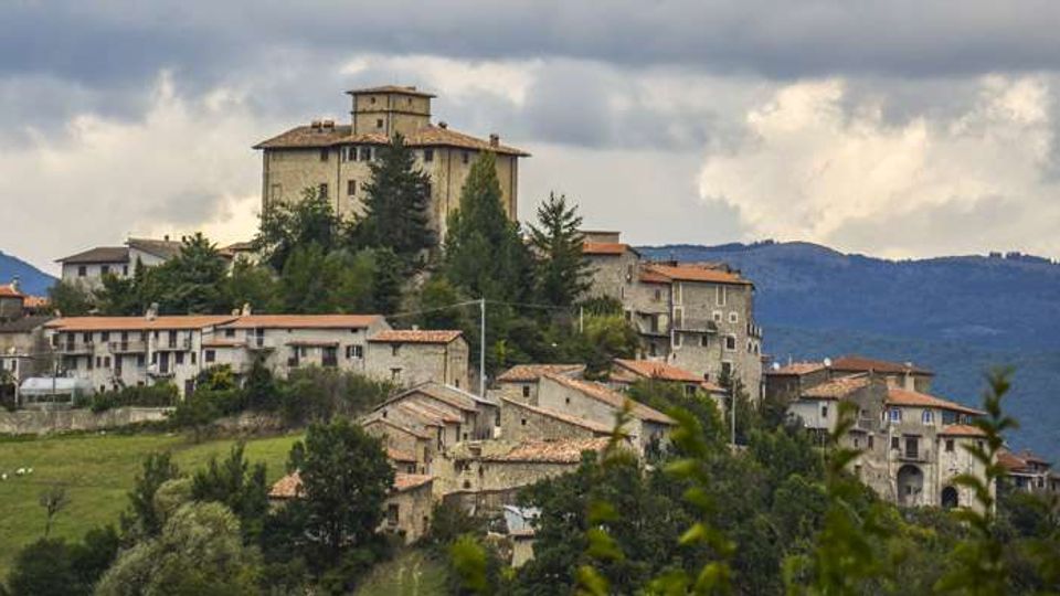 Veduta panoramica di un borgo collinare con edifici in pietra e tetti in coppi, dominato da un grande palazzo o castello su un’altura circondata da alberi. Sullo sfondo si vedono colline boscose e un cielo nuvoloso.