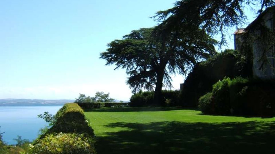 Giardino con ampio prato verde affacciato su un lago, delimitato da una siepe lungo il bordo. Grandi alberi sempreverdi proiettano ombre sul terreno e, a destra, si intravede il lato di un edificio tra la vegetazione sotto un cielo sereno.