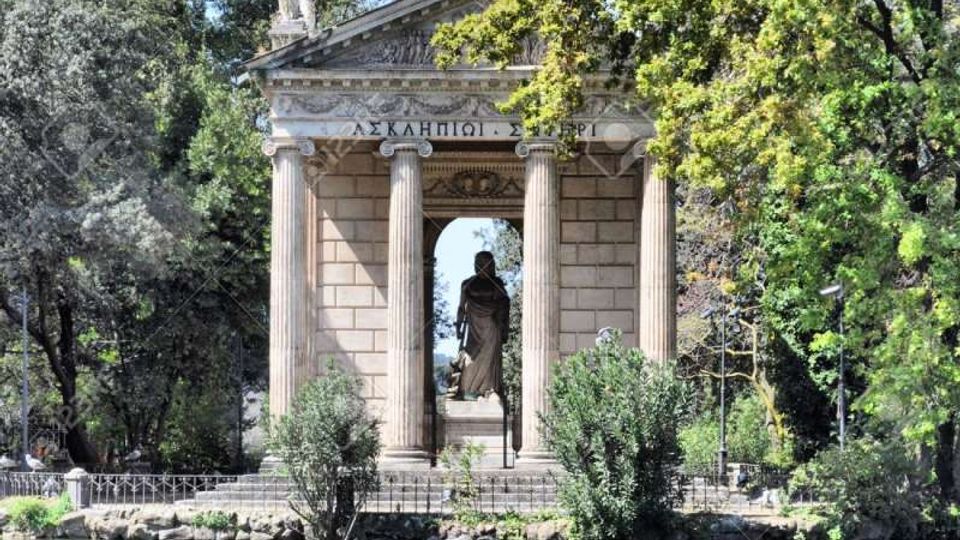 Tempietto neoclassico con frontone triangolare e colonne, con statue sul tetto e una grande scultura al centro dell’apertura. La struttura si trova sulla riva di un laghetto in un parco alberato, con vegetazione e riflessi sull’acqua in primo piano.