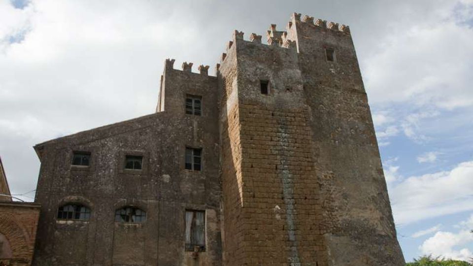 Castello in pietra con alte torri quadrate merlate e mura massicce, con finestre piccole e facciata in parte intonacata. L’edificio è ripreso dal basso contro un cielo nuvoloso, con vegetazione visibile sullo sfondo laterale.