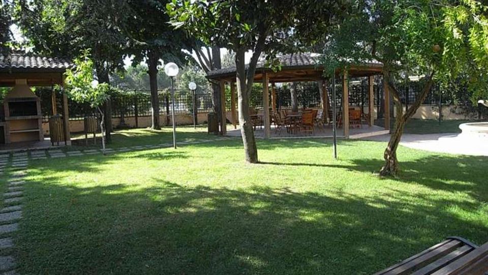 Giardino con ampio prato e alberi ombreggianti, con una panchina in primo piano e lampioni lungo il perimetro recintato. Sullo sfondo si vedono due strutture coperte in legno e tegole, una con area barbecue e l’altra con tavoli e sedie.