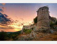 Rovine di un castello medievale su una collina rocciosa, con una grande torre cilindrica in pietra in primo piano e tratti di mura sullo sfondo. Il paesaggio circostante è collinare e boscoso, illuminato da un cielo al tramonto con nuvole sparse.