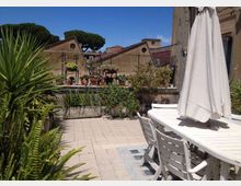 Terrazza pavimentata con tavolo e sedie bianche e un grande ombrellone chiuso, circondata da fioriere e piante in vaso lungo una ringhiera. Sullo sfondo si vedono tetti di edifici e alberi ad alto fusto sotto un cielo sereno.