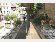 Pensilina di una fermata del tram lungo un marciapiede stretto, con binari dismessi ai lati e traversine tra ghiaia bianca. Sullo sfondo si vedono edifici urbani, vegetazione spontanea vicino ai binari e un muro con graffiti.