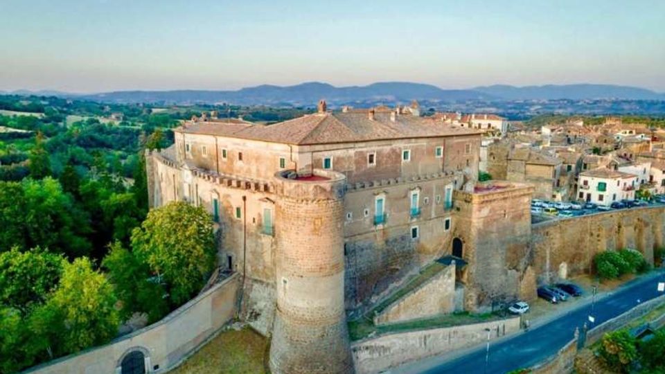 Veduta aerea di un grande castello in pietra con mura merlate e torri cilindriche, circondato da una cinta muraria e affacciato su una strada. Sullo sfondo si vede un borgo con edifici in pietra e un paesaggio collinare verde fino alle montagne all’orizzonte.