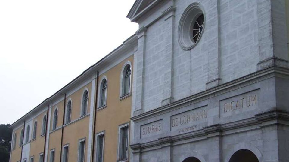 Facciata di un edificio religioso in pietra chiara con frontone e croce sulla sommità, rosone circolare e portico ad archi al piano terra con iscrizioni in rilievo. A sinistra si estende un’ala adiacente intonacata gialla con finestre ad arco, affacciata su una strada con auto parcheggiate.