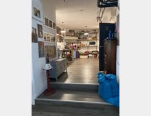 Interno di uno studio creativo con pavimento in legno grigio e un piccolo gradino d’ingresso; pareti bianche con foto e stampe, scaffali a vista pieni di materiali e un’area di lavoro illuminata da lampade a sospensione. Sul lato destro si vede una porta scura e un armadio, mentre in primo piano ci sono un pouf blu e un piccolo supporto rosso.