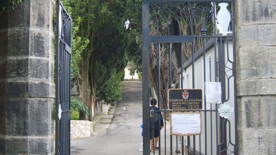Ingresso di un cimitero con cancello in ferro battuto tra pilastri in pietra, sormontato da una croce e dalla scritta “TRANSIERUNT”. Oltre il cancello si vede un viale in salita alberato, con un edificio chiaro sul lato destro e cartelli informativi affissi alla grata.