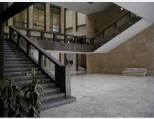 Atrio interno di un edificio con ampie scale in pietra e ringhiere scure, pavimento in marmo chiaro e pareti rivestite in lastre di pietra. Sullo sfondo si vedono grandi finestre a griglia e un ballatoio che corre lungo il piano superiore.