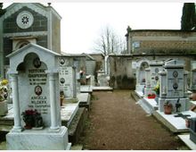 Viale centrale di un cimitero con tombe in marmo e cappelle funerarie disposte ai lati, con vasi di fiori e lastre commemorative. Sullo sfondo si vedono un edificio in muratura con facciata decorata e un alto muro perimetrale con alberi.