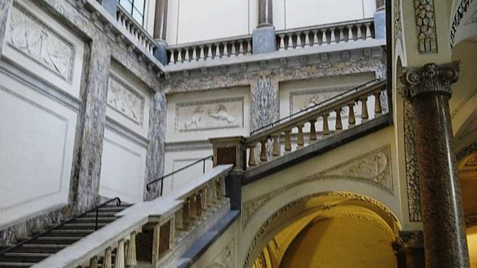 Interno di un edificio storico con ampio vano scala monumentale, pareti decorate e colonne in marmo. Grandi finestre ad arco illuminano una balconata superiore e una serie di archi e volte sottostanti.